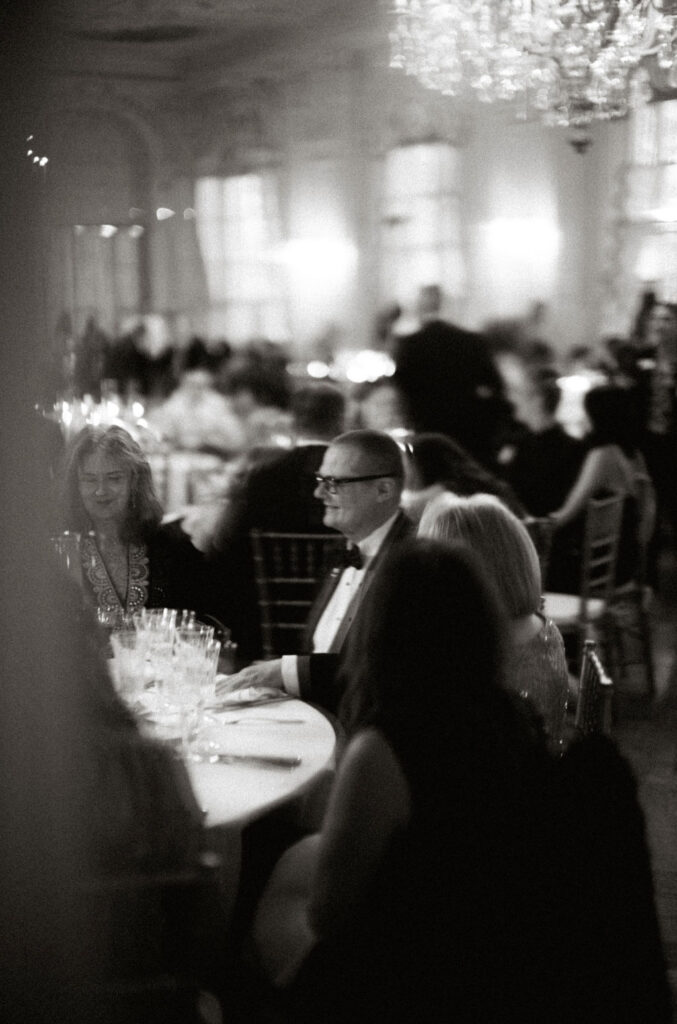 A black and white image of an older gentleman smiling at the guests at his table at a wedding. This photo is an example of how luxury wedding photographers hide in plain sight to achieve great photographic results.