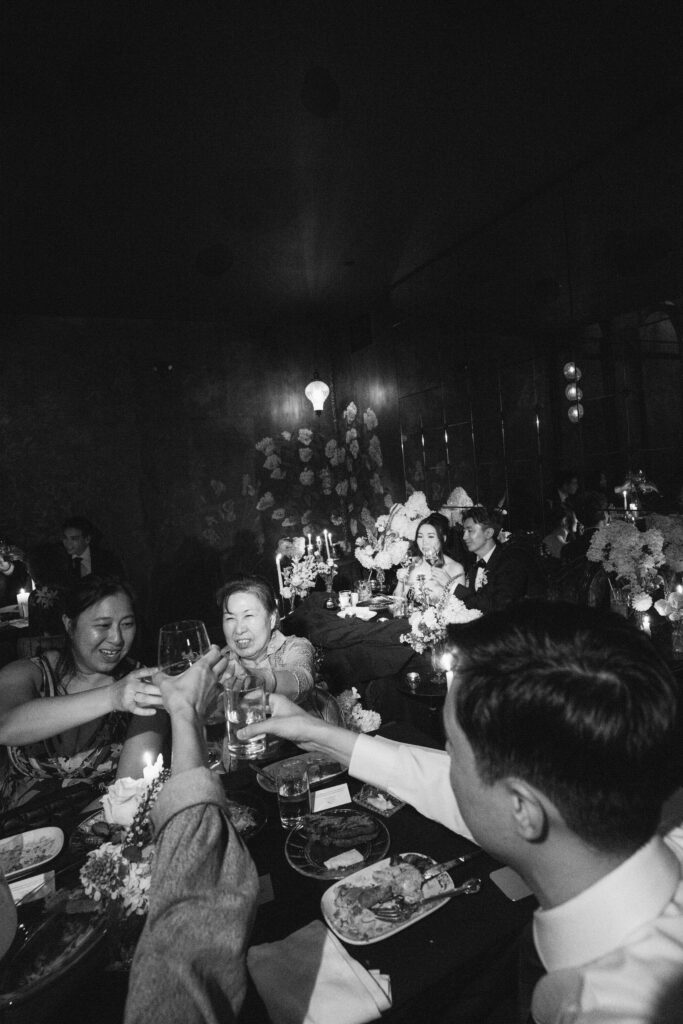 A black and white image of wedding guests clanking glasses to toast the happy couple. This photo is an example of how luxury wedding photographers hide in plain sight to achieve great photographic results.